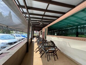 Un'area salotto esterna coperta con tavoli e sedie, una fila di auto parcheggiate e un edificio con ampie vetrate.