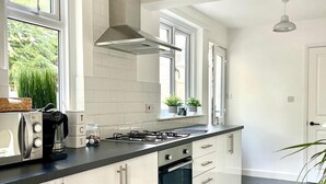 Una cocina moderna con armarios blancos, estufa a gas, horno empotrado y campana extractora de acero inoxidable. Hay ventanas que ofrecen luz natural y vistas a zonas verdes.