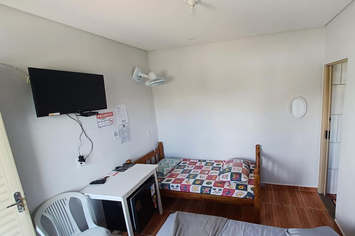 Une petite pièce, peu meublée, comprenant un lit, un bureau, une chaise, un téléviseur fixé au mur et une porte.