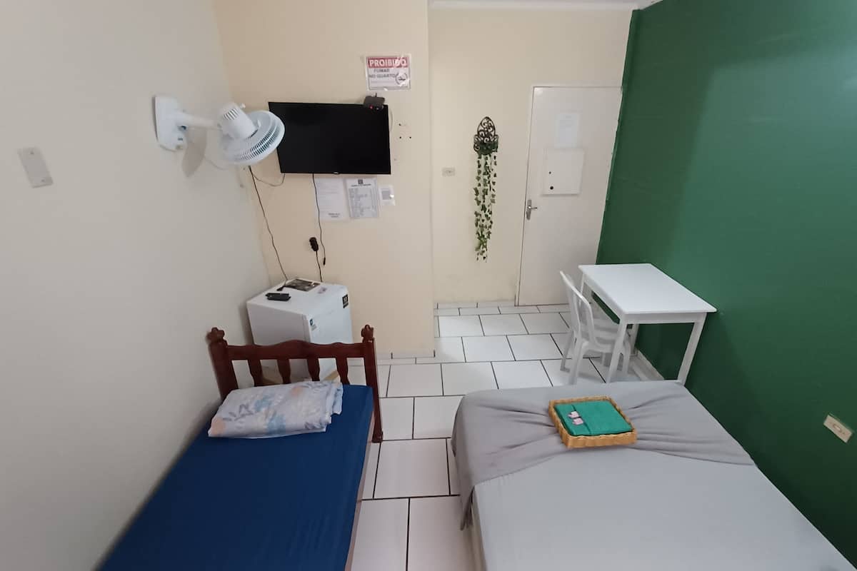 Une petite pièce comprenant un lit, un bureau, une chaise, un ventilateur fixé au mur et un téléviseur.