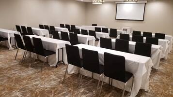 Un salón de conferencias con mesas y sillas dispuestas en filas, una pantalla de proyector y una alfombra estampada.