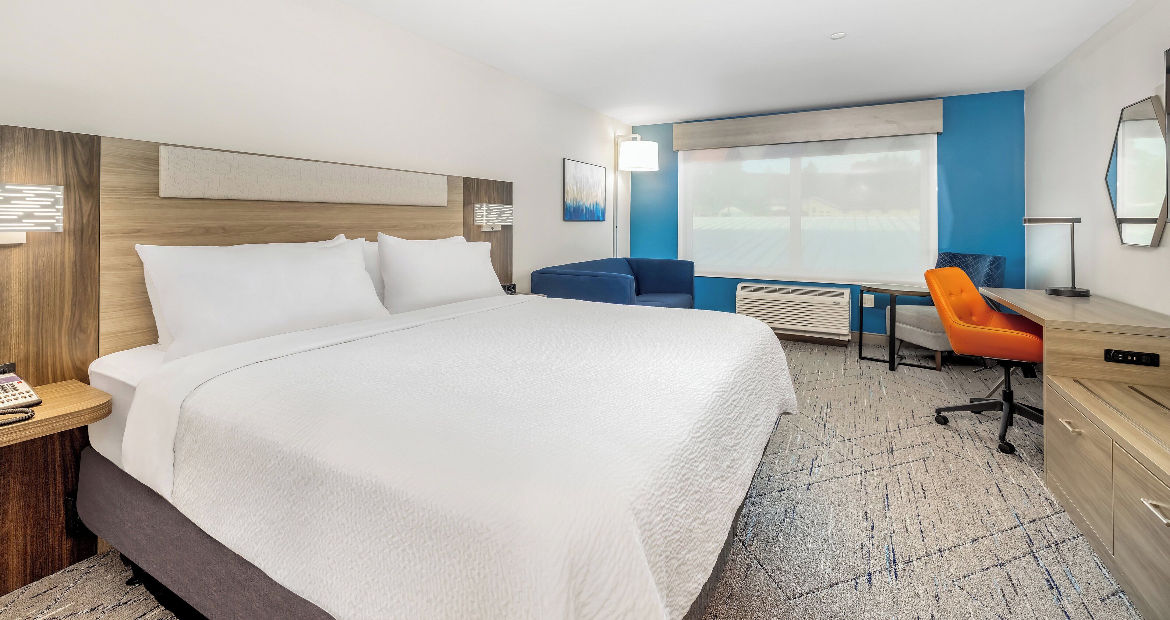 A hotel room with a large bed, a desk with a chair, a window, and a wall-mounted air conditioner.