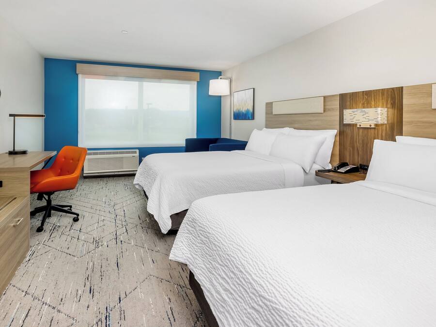 A hotel room with two beds, a desk, an orange chair, a large window, and a wall-mounted TV.