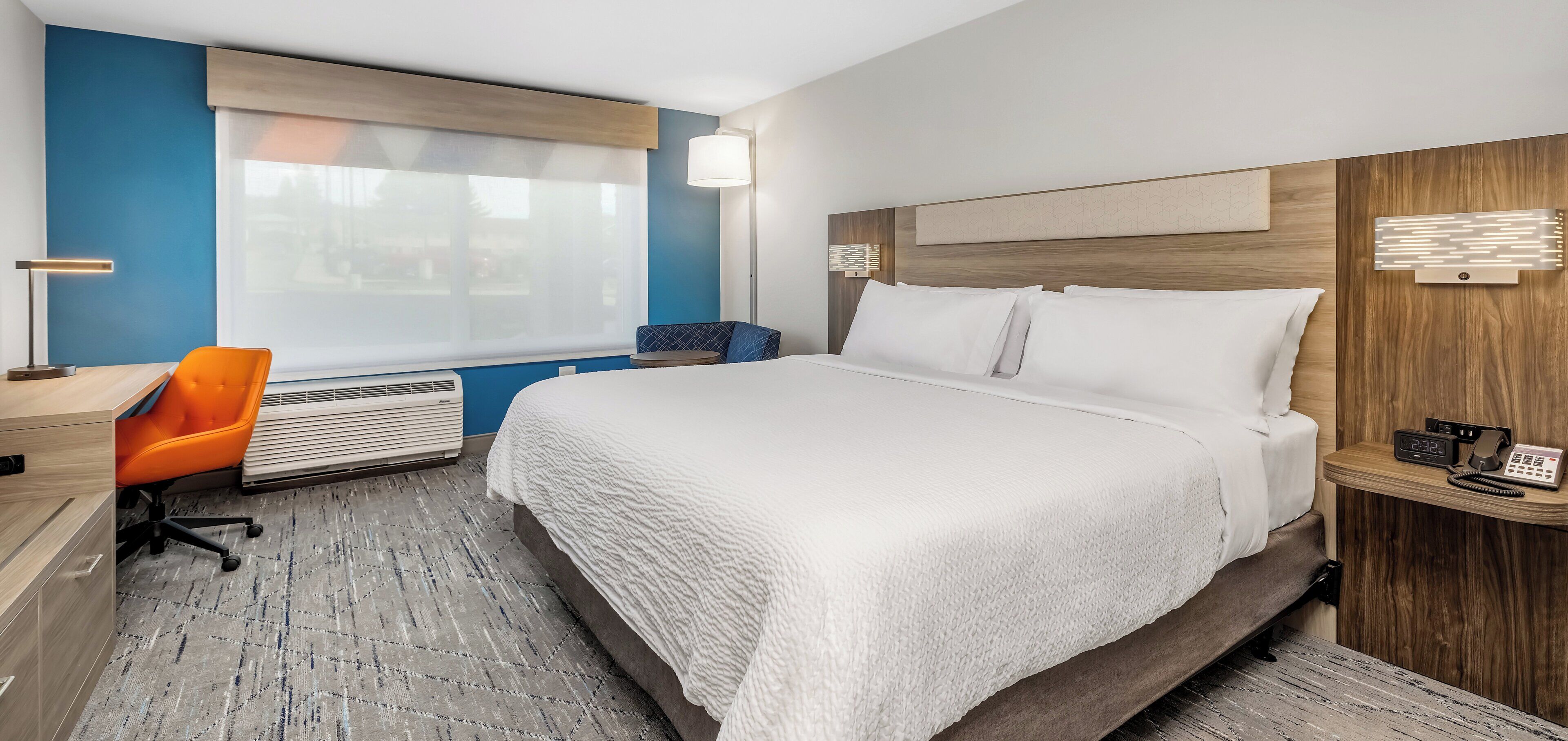 A hotel room with a large bed, a desk with a chair, a bedside table with a phone, a wall-mounted lamp, and a window with blinds.