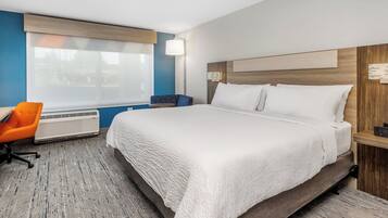 A hotel room with a large bed, a desk with a chair, a bedside table with a phone, a wall-mounted lamp, and a window with blinds.