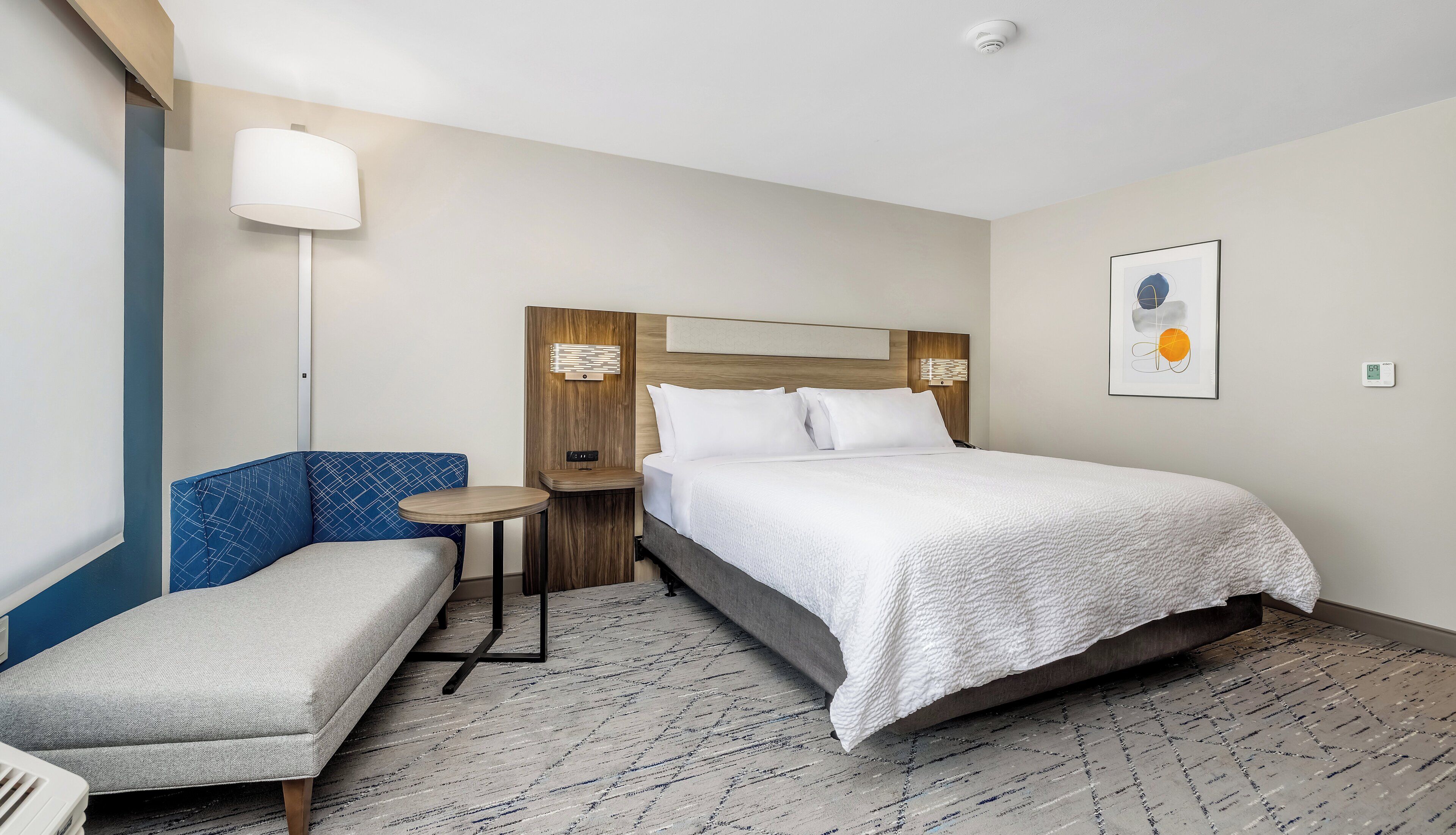 Una habitación de hotel con una cama, un banco, una mesita, una lámpara y un cuadro en la pared.