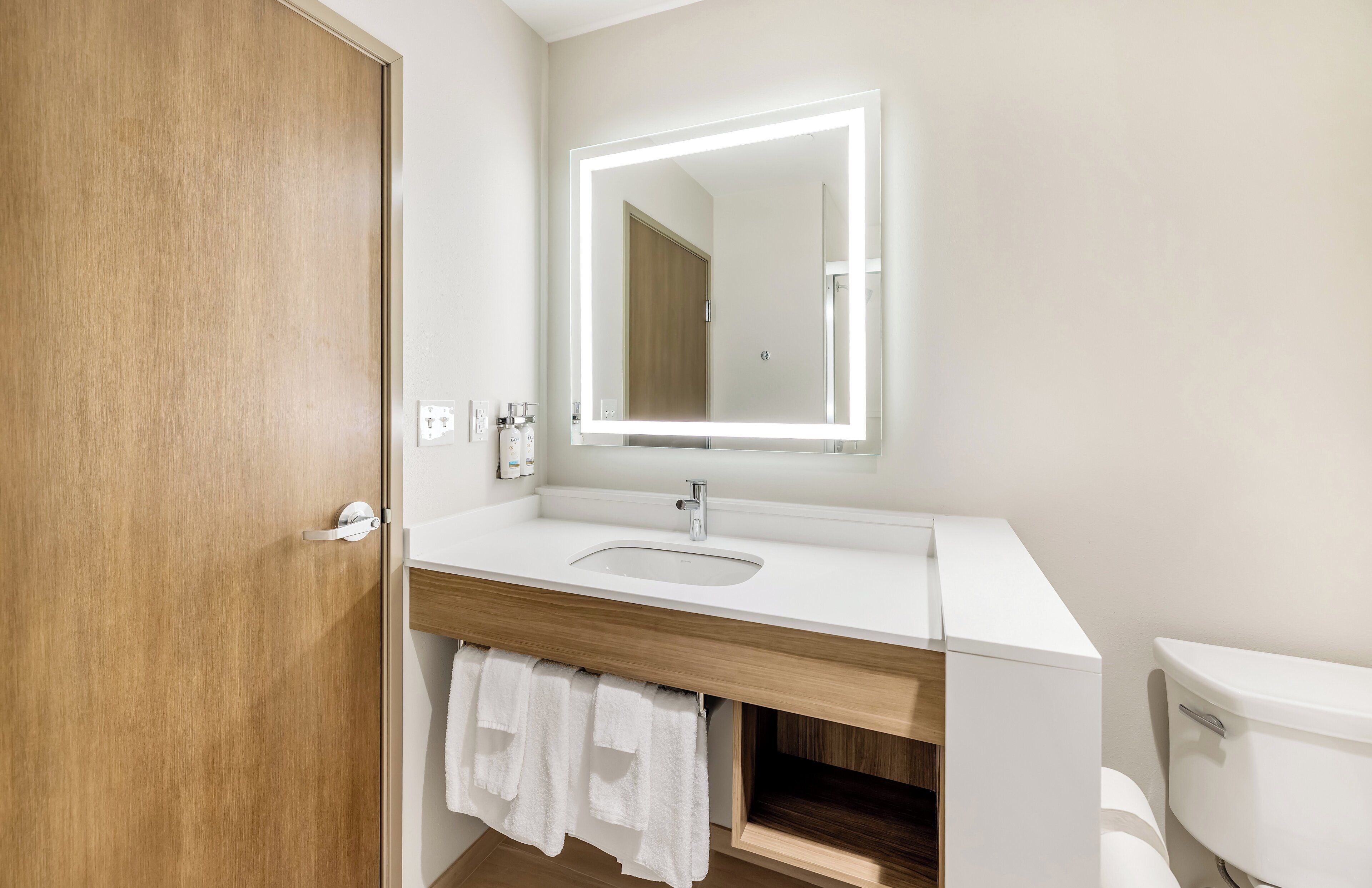 Ein Badezimmer mit einem Holzbadezimmerhocker, einem weißen Waschbecken, einem Spiegel mit Beleuchtung, einer Toilette und einem Handtuchhalter.