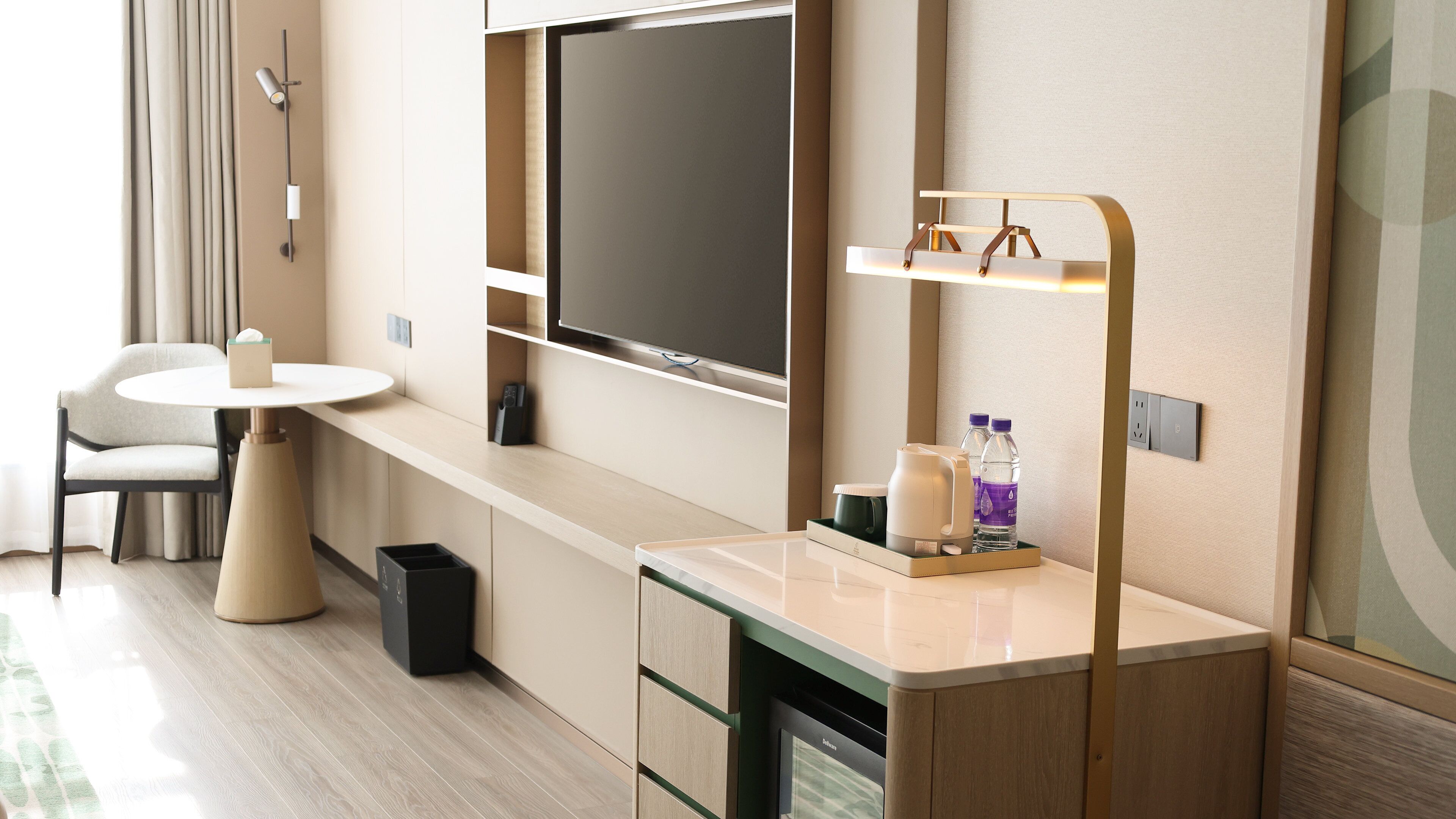 Une chambre d’hôtel moderne équipée d’une télévision à écran plat, d’une petite table avec une chaise, d’une table de chevet avec une lampe et d’une bouteille d’eau.