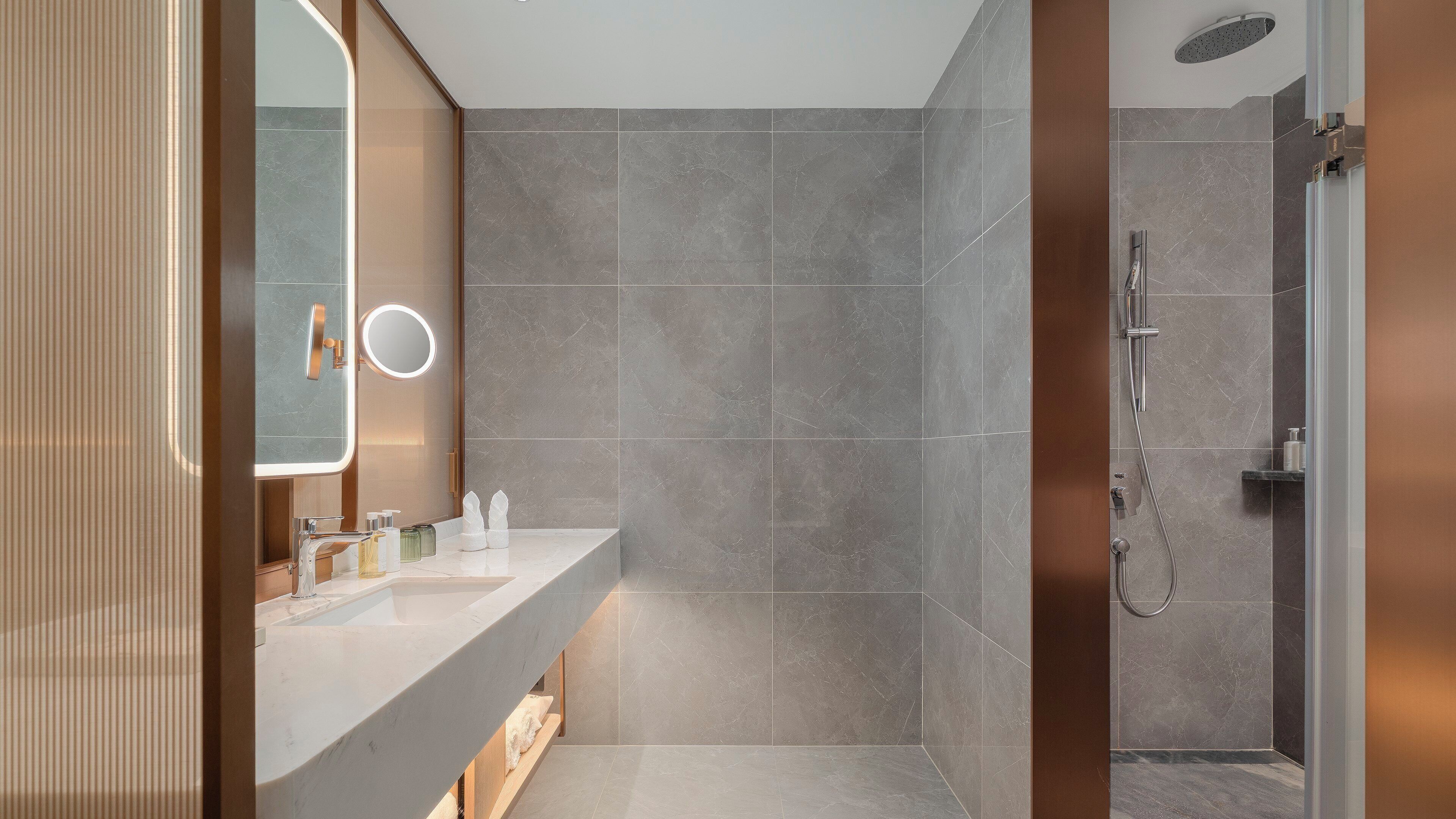 A modern bathroom with a large mirror, a round light, a shower, and a vanity with toiletries.