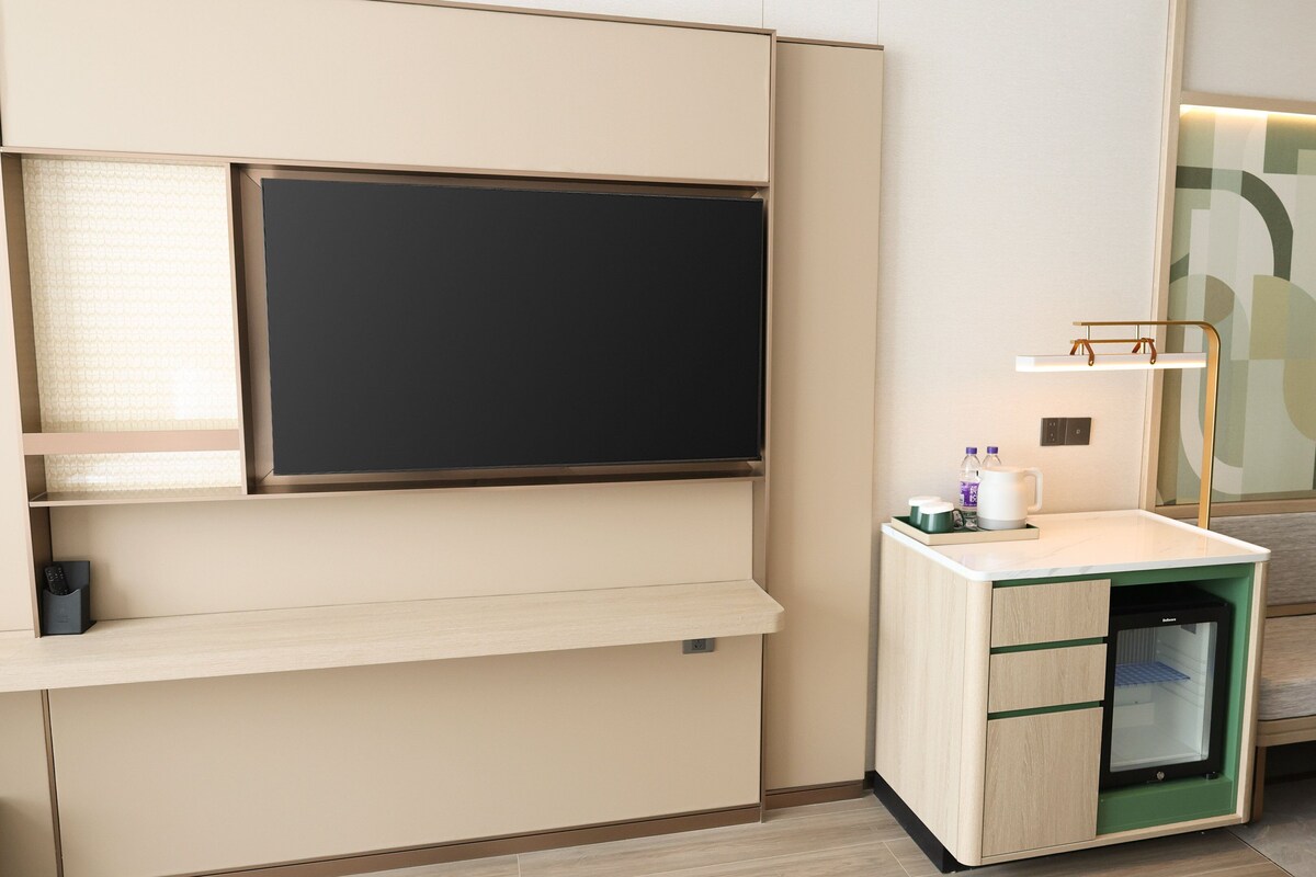 A modern hotel room with a built-in TV, a small kitchenette, a seating area, and a large mirror.