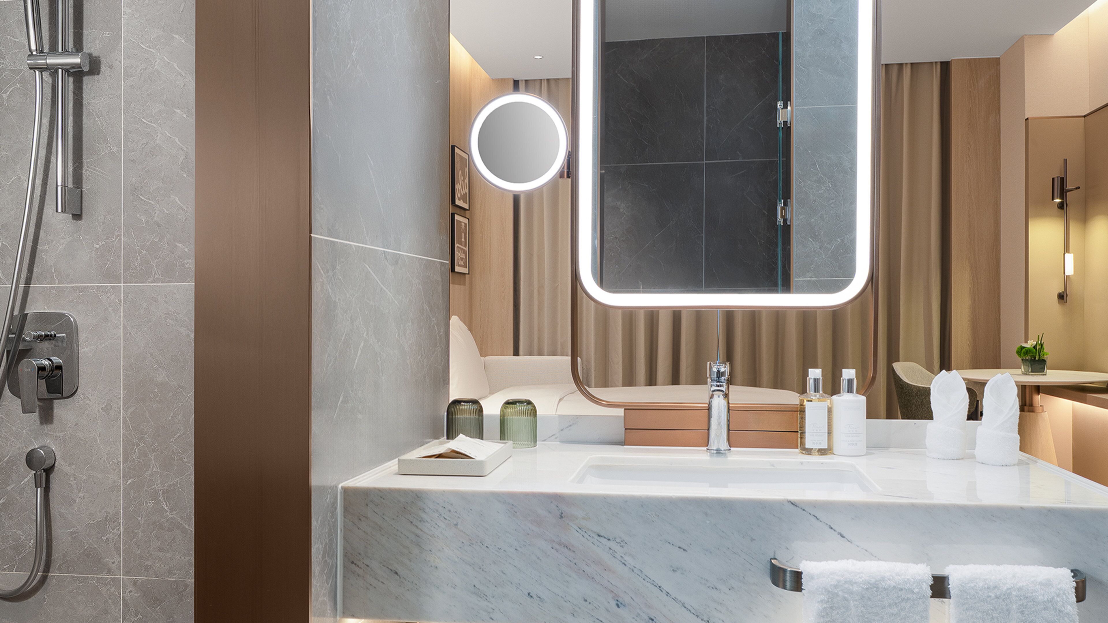 A modern bathroom with a marble sink, a large mirror with a light, a handheld showerhead, and a towel rack.