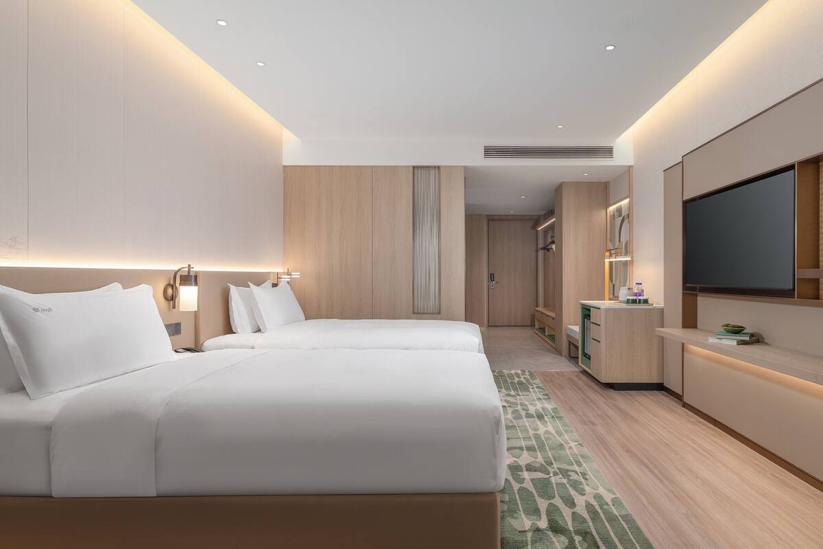 A hotel room with two beds, a desk, a TV, and a bathroom in the background.