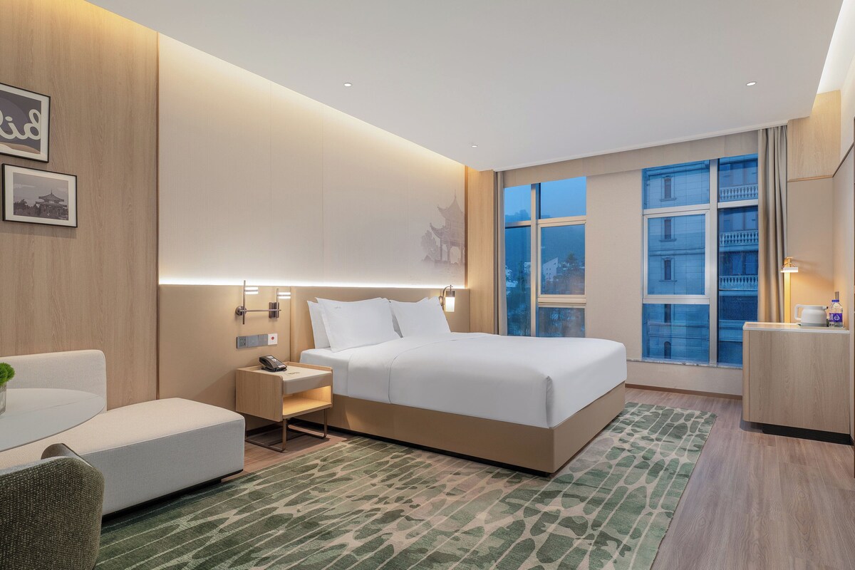 A modern hotel room with a large bed, bedside tables, a desk, and a view of the city.