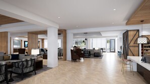 A spacious, modern living area with a herringbone wood floor, a central seating area with sofas and chairs, and a kitchen visible in the background.