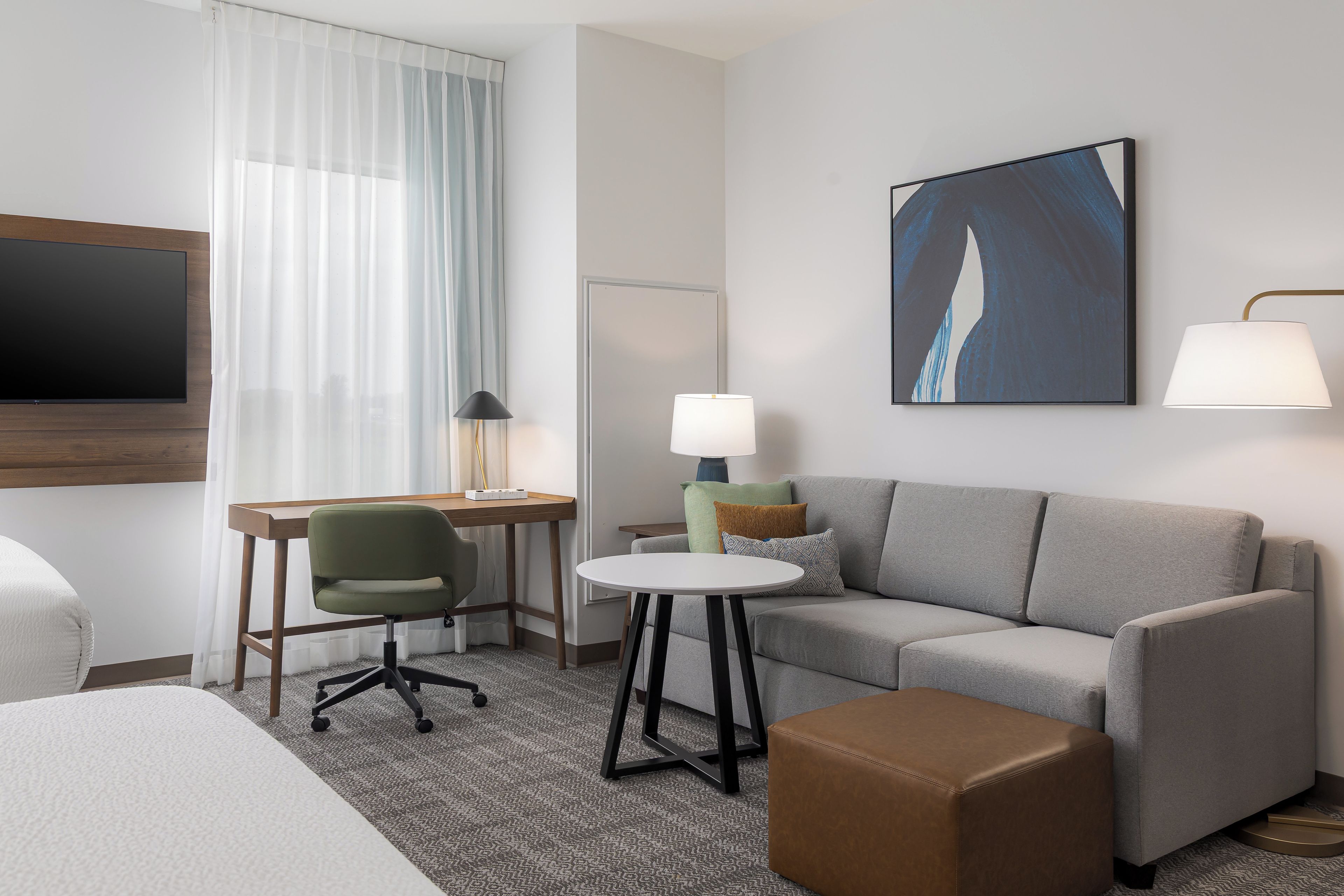A hotel room with a bed, a desk, a chair, a sofa, a coffee table, a lamp, and a painting on the wall.