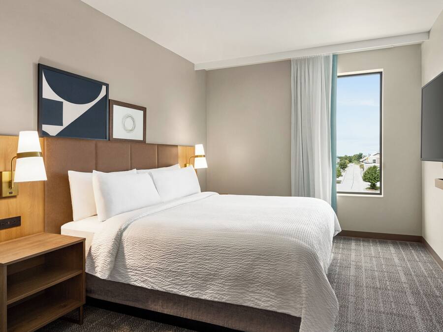 Quarto de hotel com uma cama grande, duas luminárias de cabeceira, painel de cama com interruptor de luz, televisão de tela plana, janela com vista externa e uma pequena mesa de cabeceira de madeira.