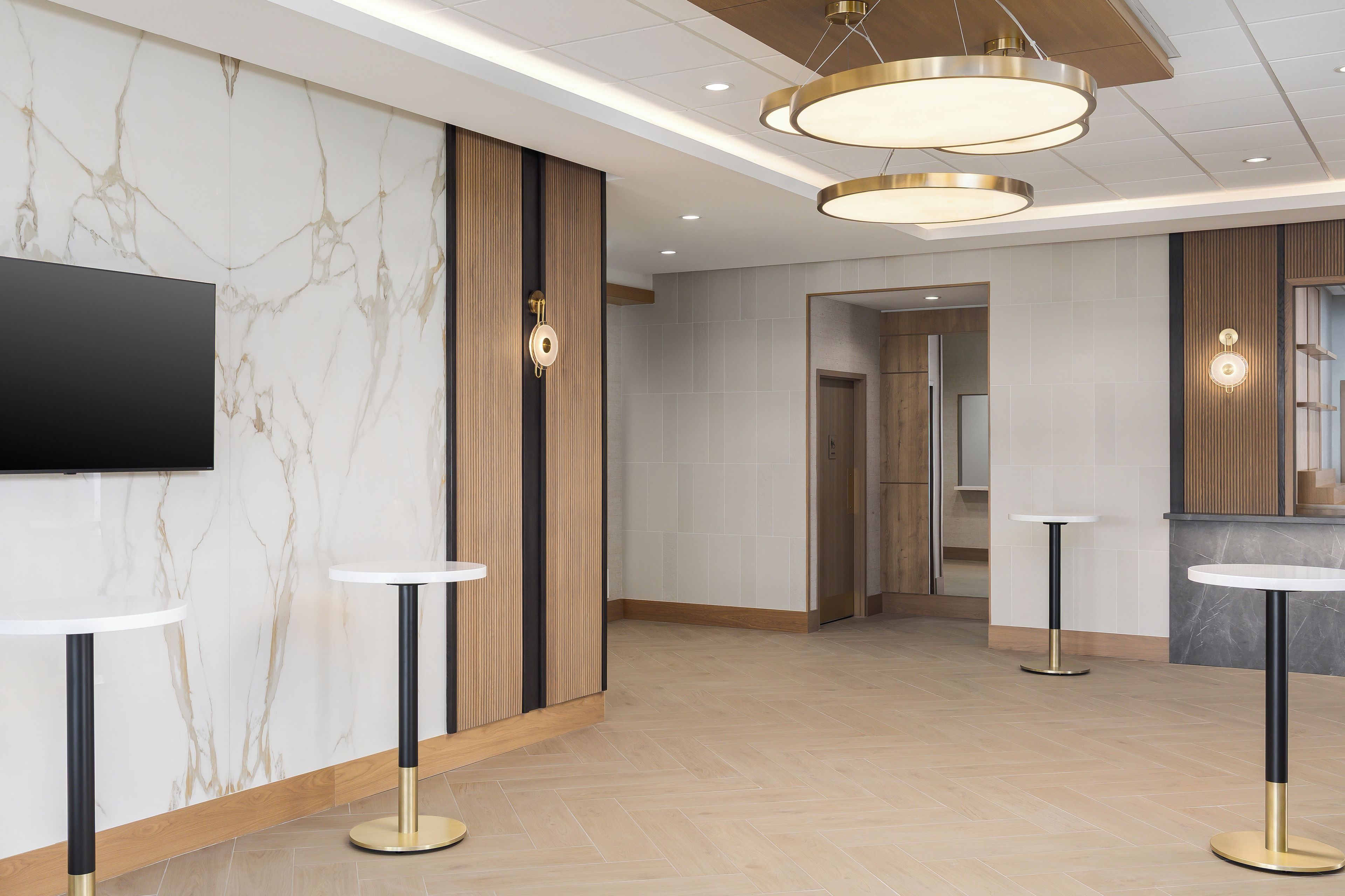 A hotel lobby with marble walls, a flat-screen TV, two round tables with black and gold bases, and a wooden reception desk.