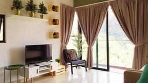 A modern living room with a flat-screen TV, a glass coffee table, a sofa, and a chair with a colorful cushion. There are potted plants on a shelf and a large window with curtains. - Electus Home at Midhills Genting - Free WiFi & Parking (Genting Highlands)