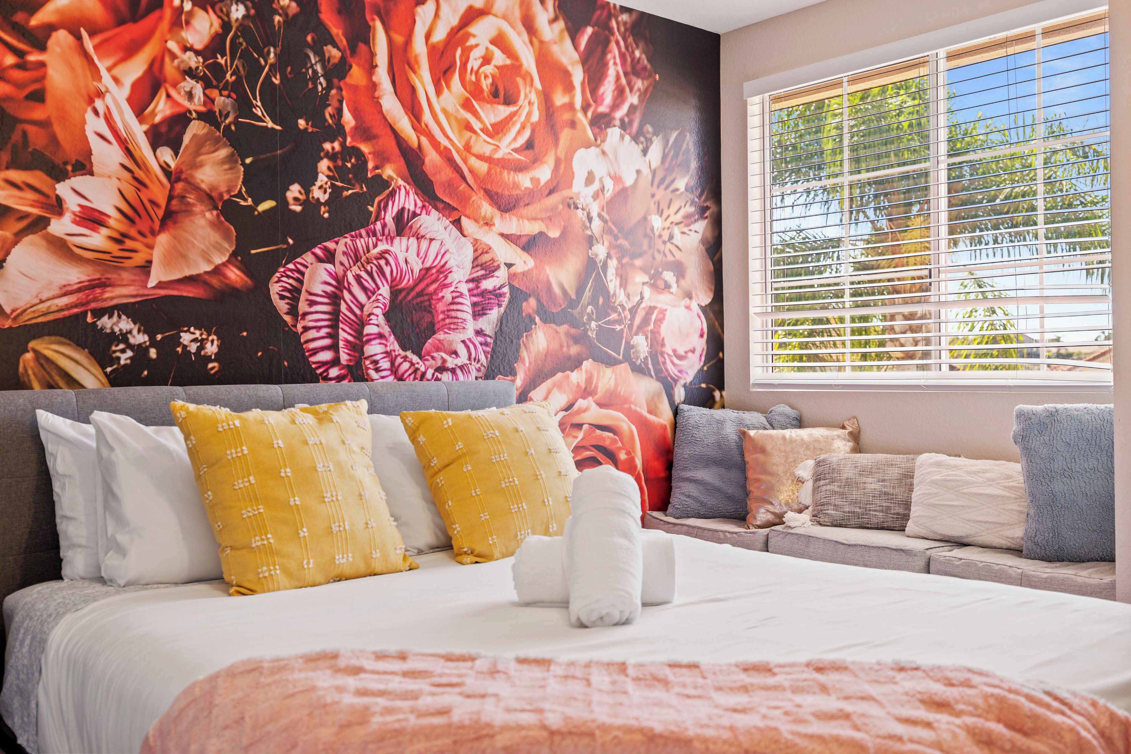 Een slaapkamer met een groot bloemenmural, een bed met witte lakens en een raam met zonwering.