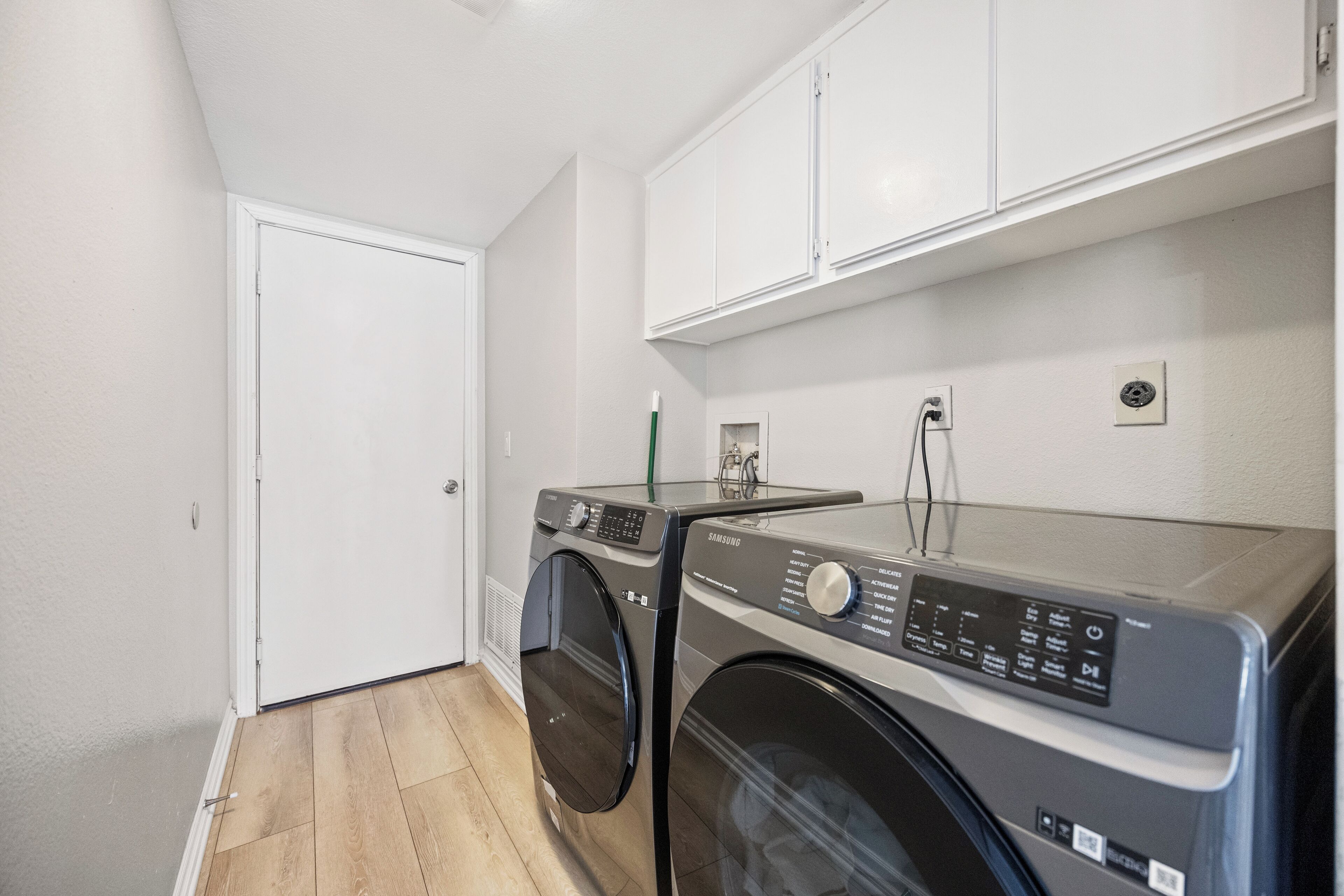 A laundry room with a front-loading washer and dryer, white cabinets above, and a door to the left.