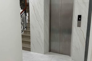 Un ascenseur dans un couloir aux murs de marbre et un escalier avec une rampe en métal.