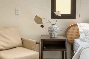 A room with a beige armchair, a bedside table with a vase, a wicker headboard, and a window above the bed. - Signy House Saigon Center - Nguyen Trai (Ho Chi Minh City)