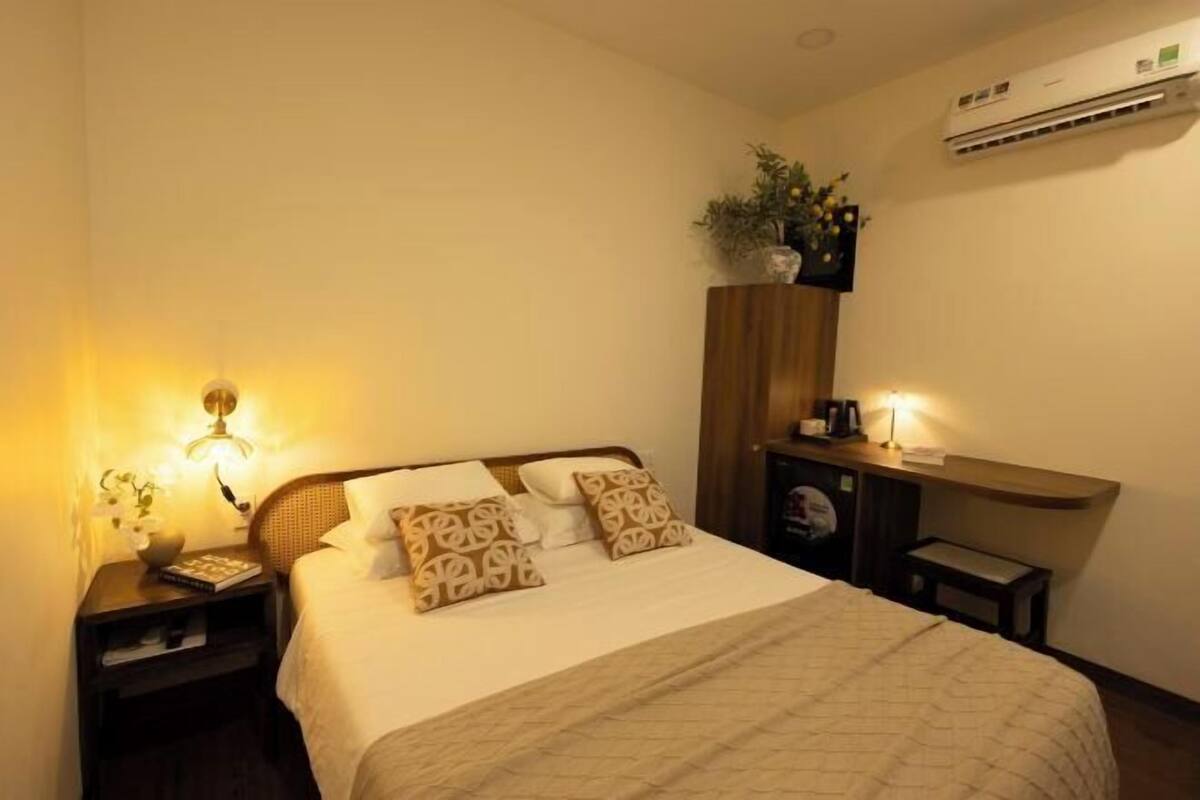 A hotel room with a bed, bedside lamps, a nightstand, a small table, a shelf with a plant, and an air conditioning unit.