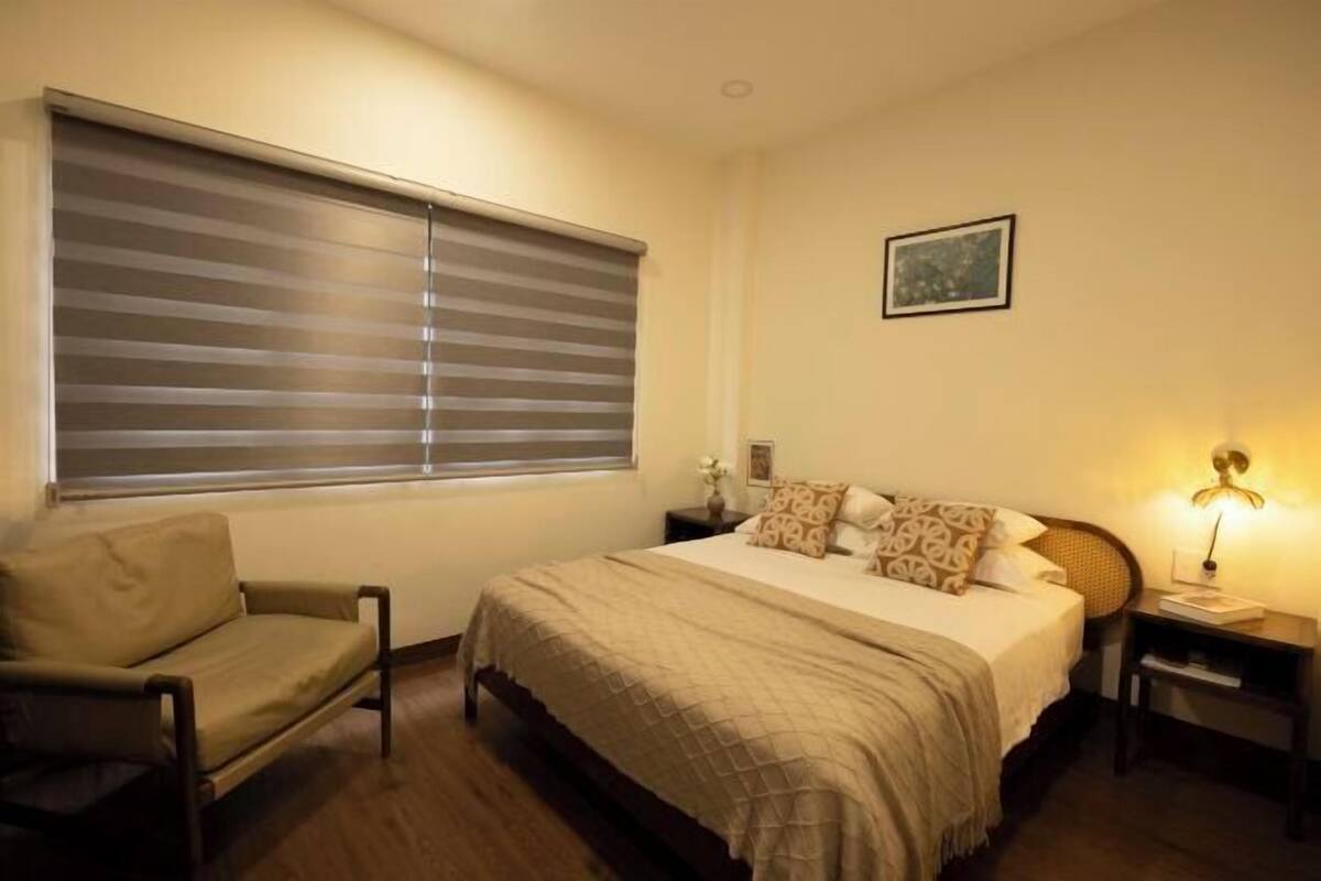 A bedroom with a bed, a chair, a bedside table, a lamp, and a window with blinds.