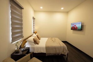 A hotel room with a bed, a TV mounted on the wall, a bedside table with a lamp, and a window with blinds. - Signy House Saigon Center - Nguyen Trai (Ho Chi Minh City)