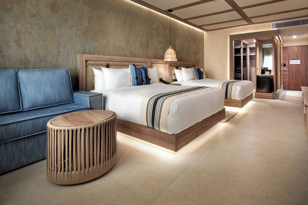 A modern hotel room with two beds, a sofa, a round wooden stool, and a walk-in closet.