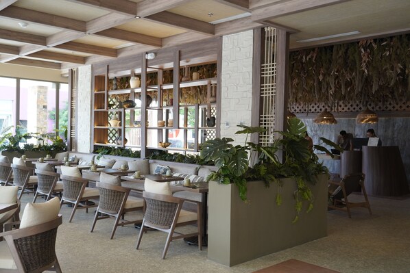 A modern interior with a long dining area, wicker chairs, and a central planter with greenery.