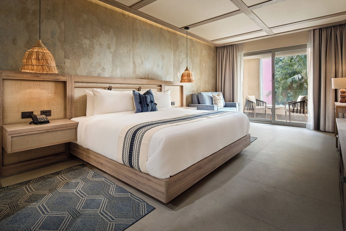 A modern hotel room with a large bed, a bedside table with a lamp, a telephone, and a window with curtains.