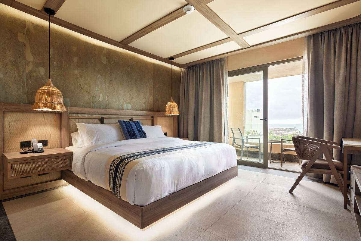 A modern hotel room with a large bed, bedside tables, a desk, and a view of the outdoors.