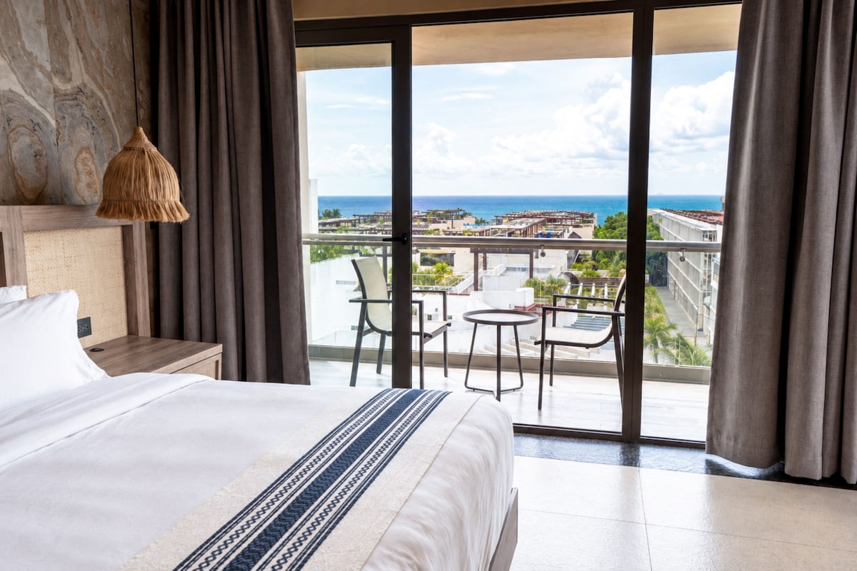 A hotel room with a large bed, a view of the sea, and a balcony with chairs and a table.