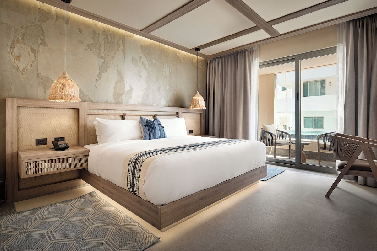 A modern hotel room with a large bed, a bedside table with a phone, a hanging light fixture, and a view of the outside balcony.