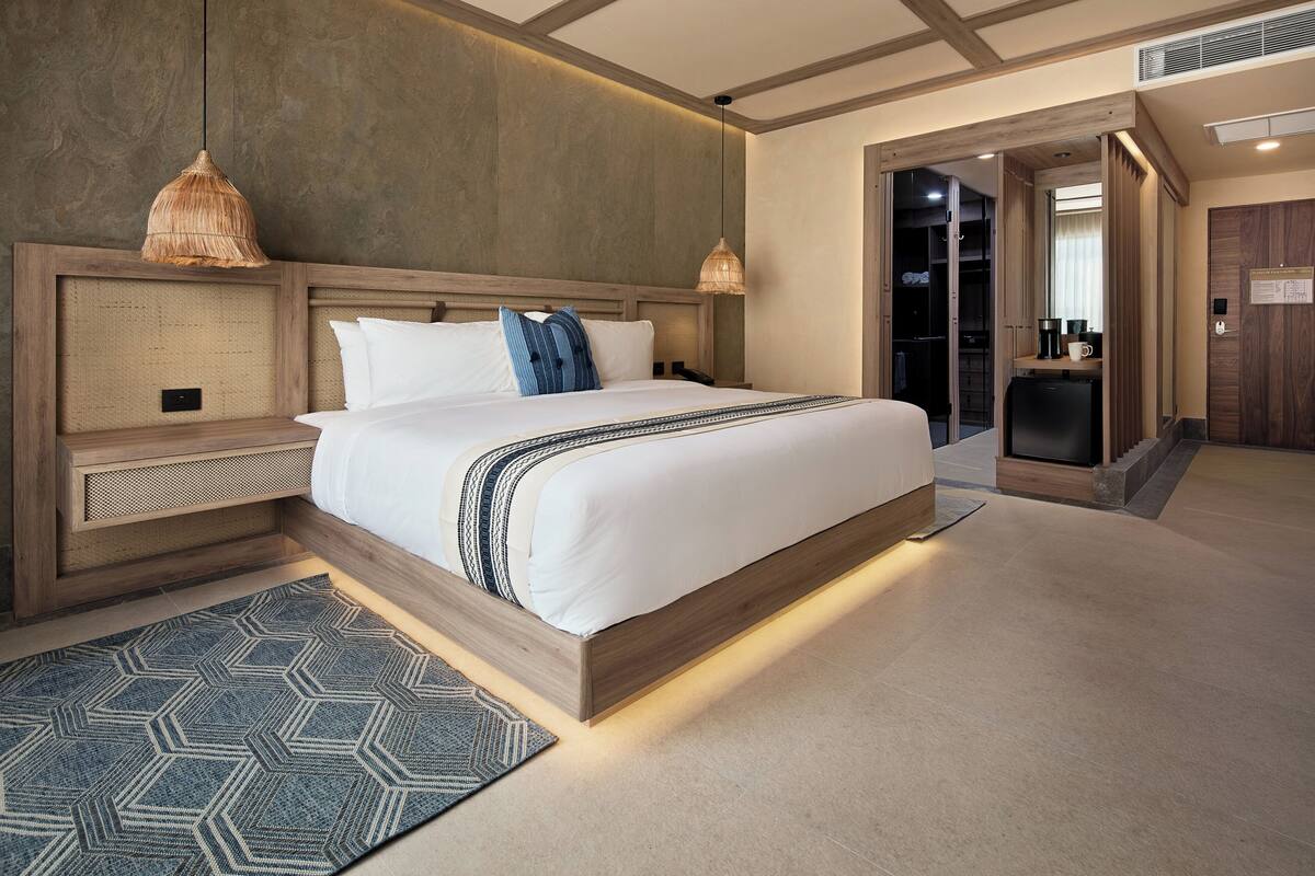 A modern hotel room with a large bed, a wooden headboard, a built-in desk, and a walk-in closet.