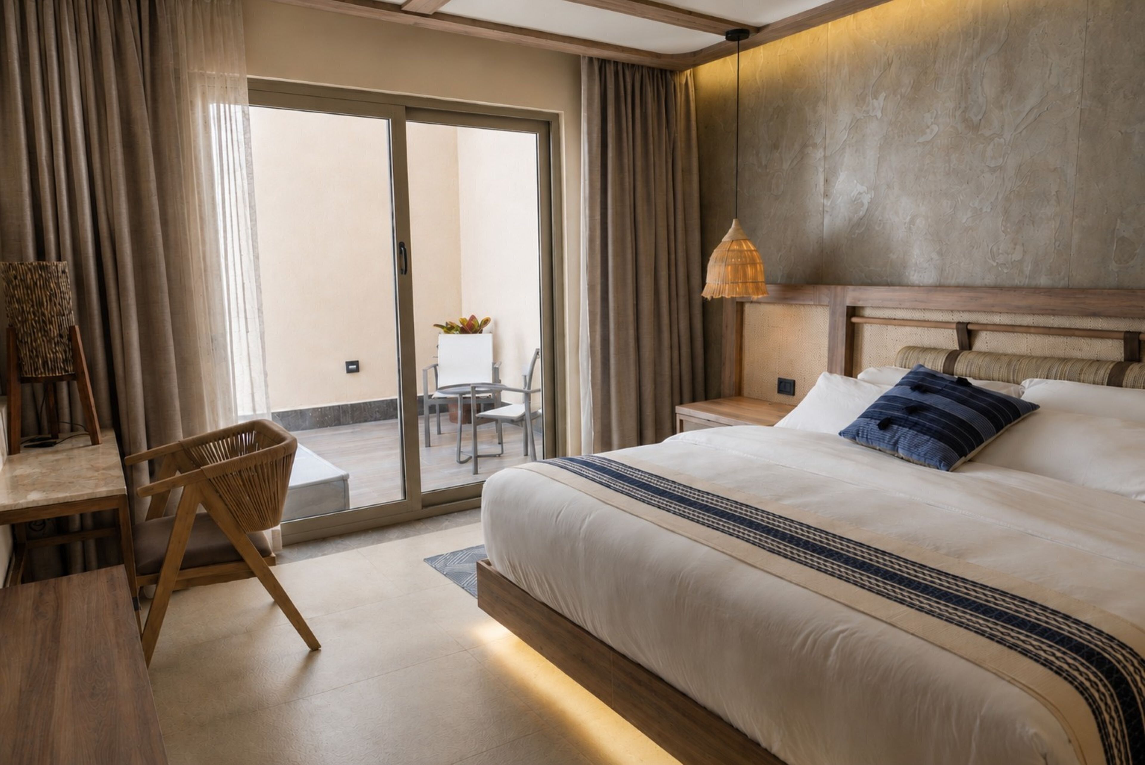 A hotel room with a large bed, a small table with chairs, a lamp, and a view through a sliding door to an outdoor area.