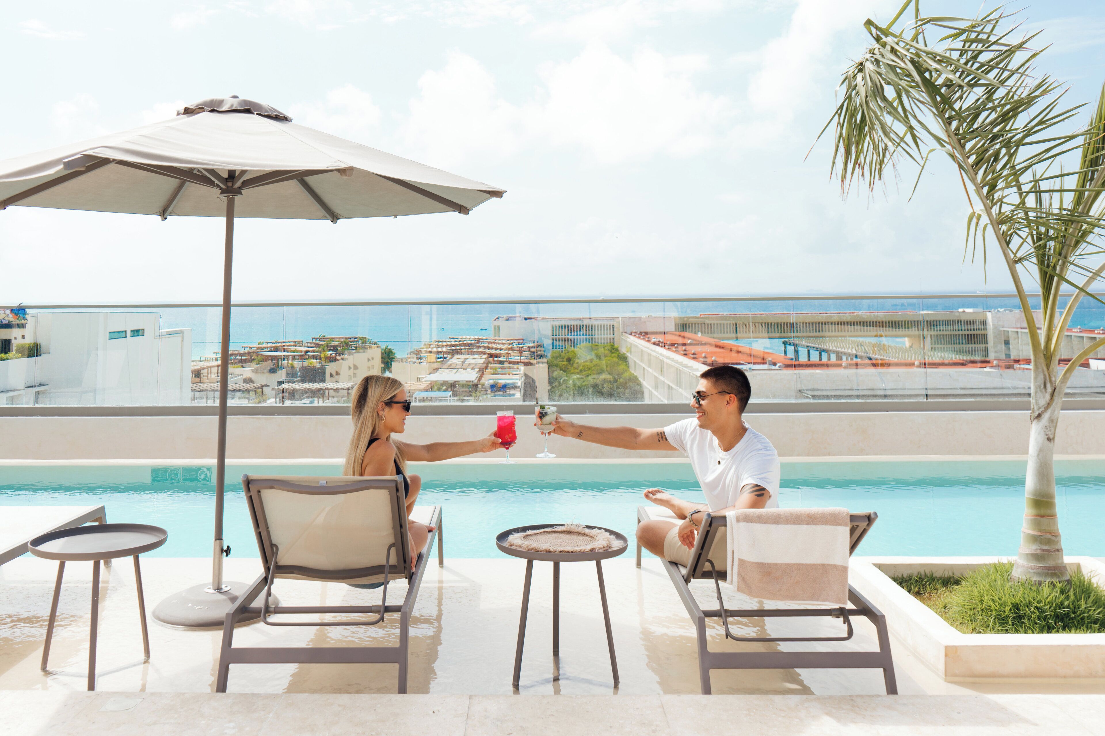 Un couple trinque avec des boissons au bord d’une piscine, avec une vue sur le paysage urbain.