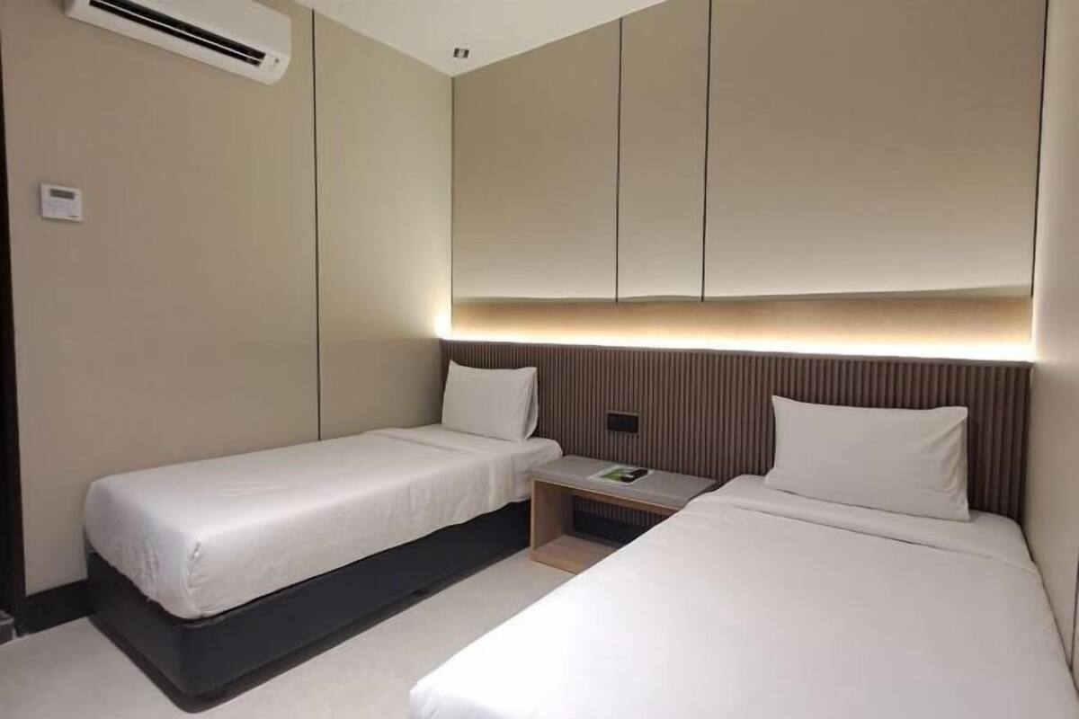 A hotel room with two single beds, a bedside table with a lamp, and an air conditioning unit on the wall.