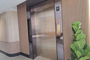 An elevator with metallic doors and a control panel, surrounded by wooden paneling and a potted plant. - Urban Inn Myra Meranti (Puchong)