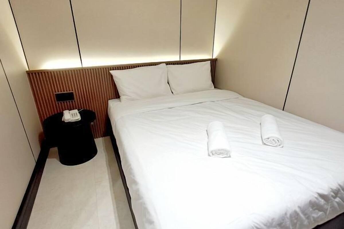 A neatly made bed with white linens and two rolled towels, a black trash bin, and a small black object on the nightstand.