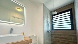 Et moderne badeværelse med bruseniche i glas, et hvidt håndvaskarrangement med træbordplade og et toilet.