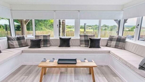 A room with a built-in bench seating area, a wooden table, and large windows offering a view of the outdoors. - Bagend Sleeps 14 Hot Tub Balcony & Ping Pong (North Devon District)