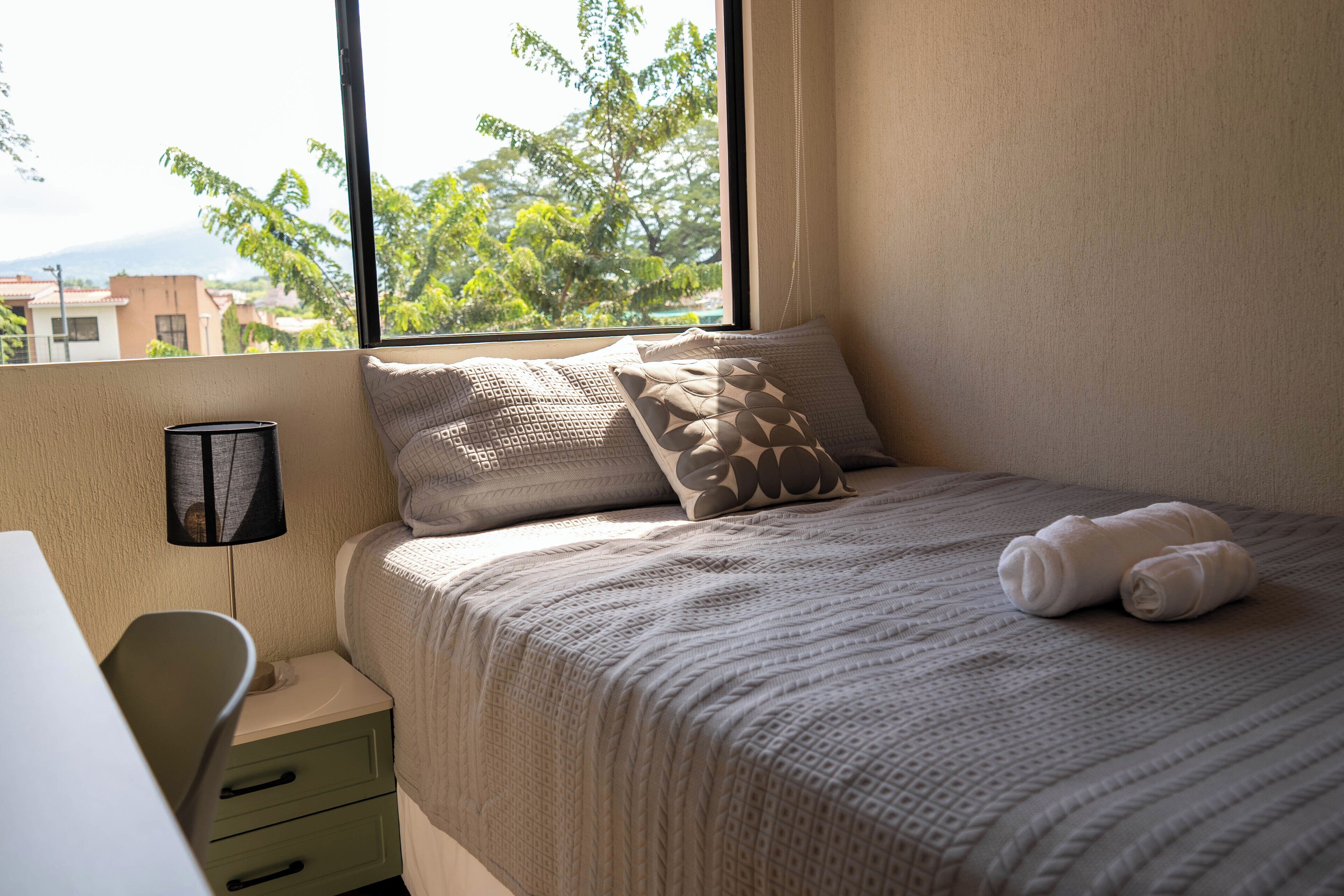 A neatly made bed with a patterned bedspread and pillows, a bedside lamp, and a view of trees and buildings through a window.