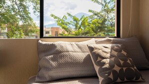 A window with a view of trees and buildings, a sofa with patterned cushions, and a curtain. - Family Oasis w/ Full A/C • Pools & 24/7 Security (Apopa)