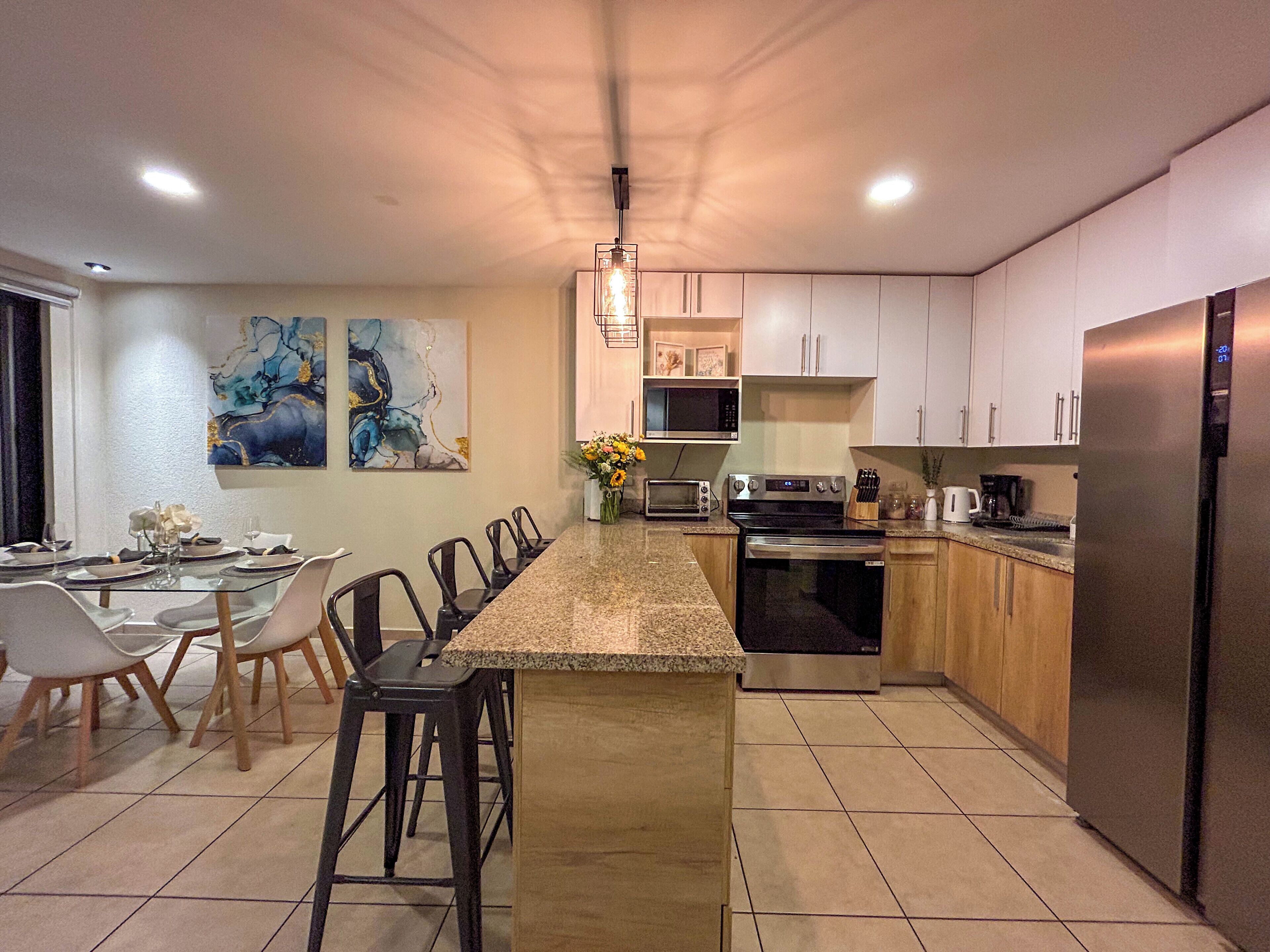 Cocina moderna con isla central, electrodomésticos de acero inoxidable y comedor con mesa y sillas de vidrio.