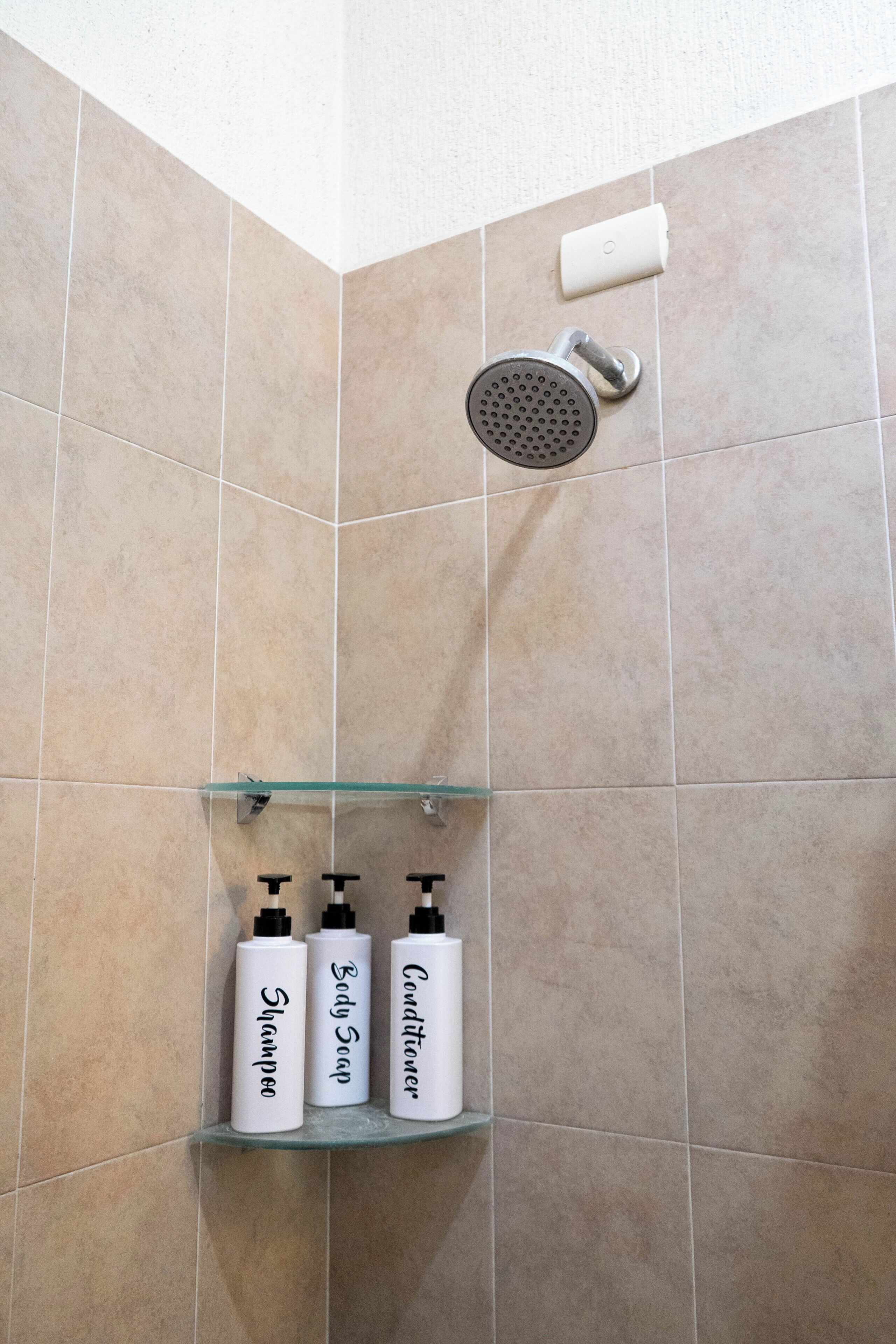 Et badeværelse med beige fliser, et brusehoved og tre flasker shampoo, sæbe og balsam på en glashylde.