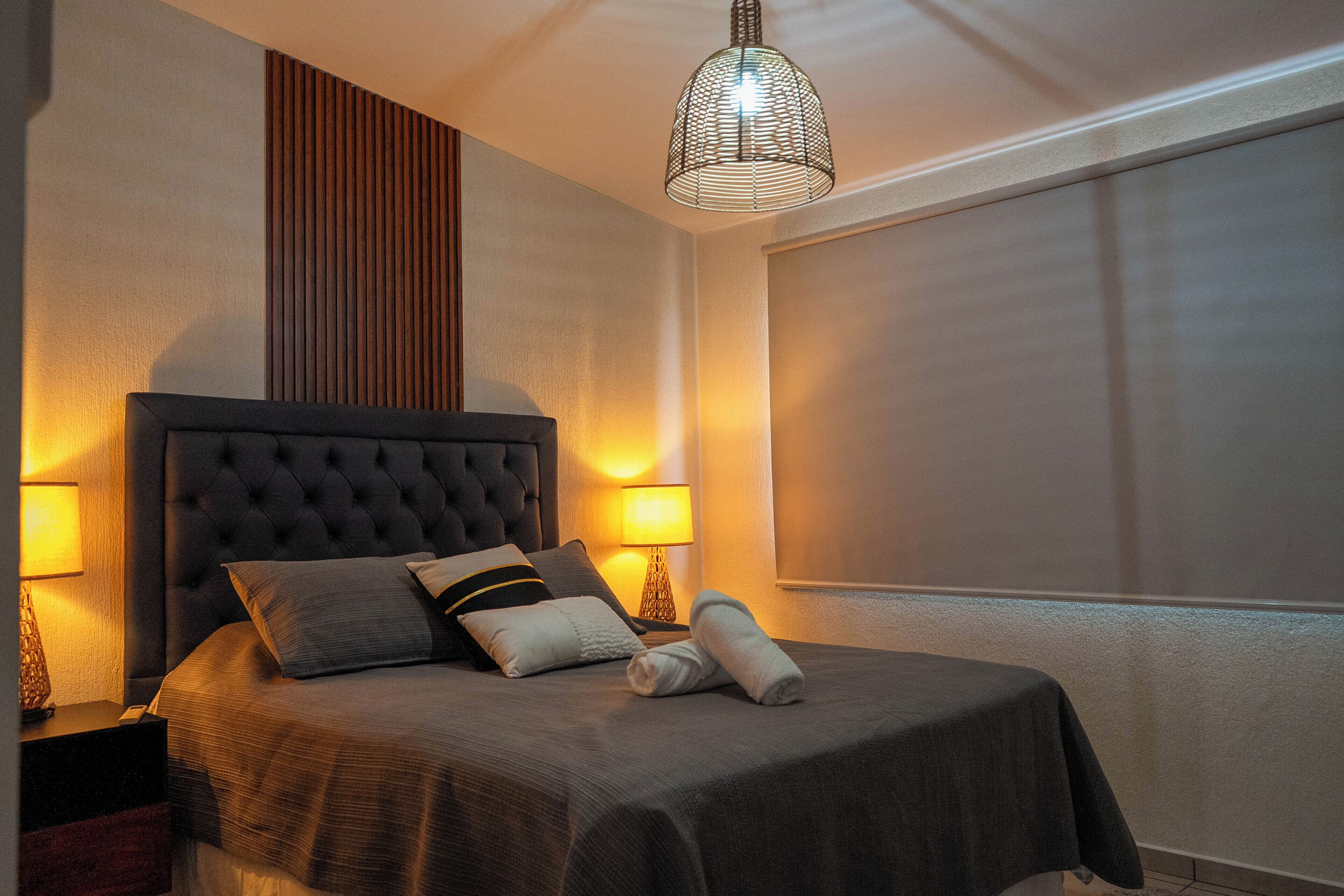 A neatly made bed with a dark headboard and pillows, a bedside lamp, and a hanging light fixture.