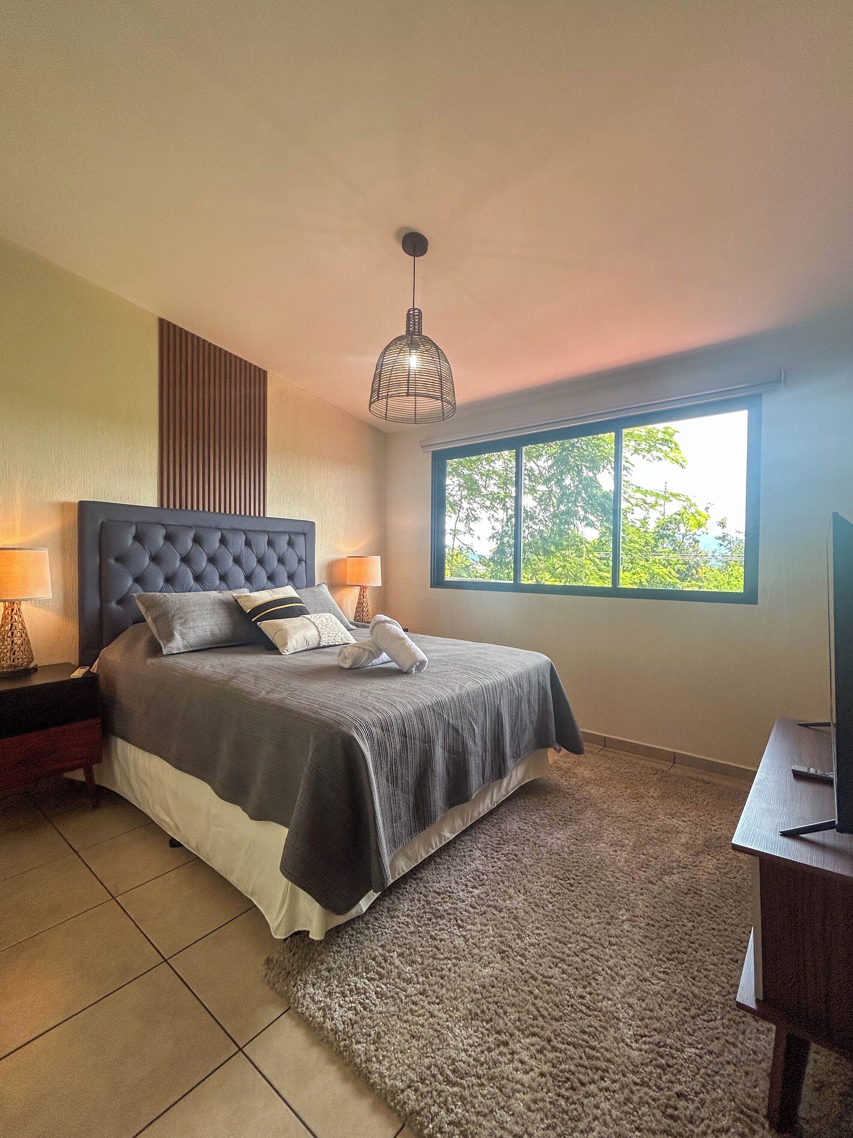 Un dormitorio con una cama grande, un televisor sobre un mueble, una ventana con vistas a zonas verdes, y una lámpara colgante.