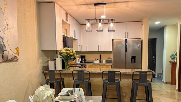 A modern kitchen with white cabinets, a stainless steel refrigerator, and a dining area with a table set for four. - Family Oasis w/ Full A/C • Pools & 24/7 Security (Apopa)